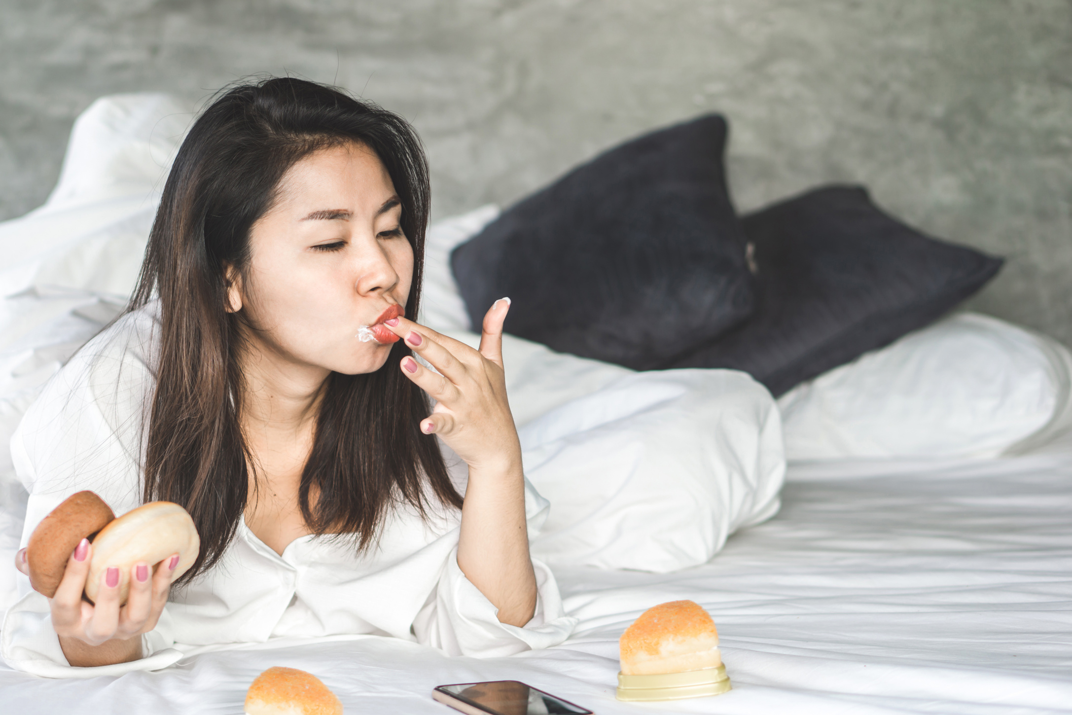 Indulging in Sweet Delights: Asian Woman Enjoying Desserts in Bed, Reflecting on the Concept of Overeating Sugar
